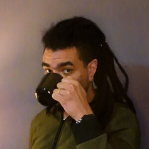 Um homem pardo com locks tomando café com uma xícara preta. Ele veste um casaco verde e tem uma parede cinza atrás dele.